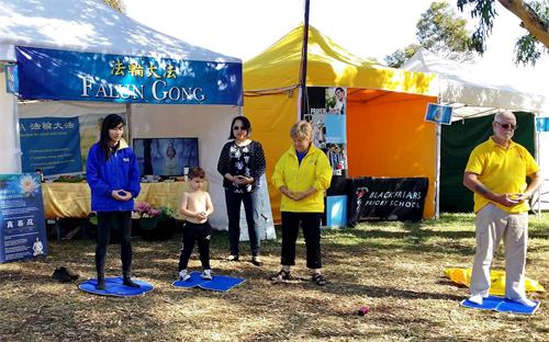 '图5:图中男孩只有四岁,因为玩热了,他母亲帮他脱下上衣。也许感受到功法展示的祥和能量场,他径直跑到法轮功展位来,拿起一个坐垫,站到正在展示功法的学员当中,认真跟着学起炼功动作,样子纯真可爱。'