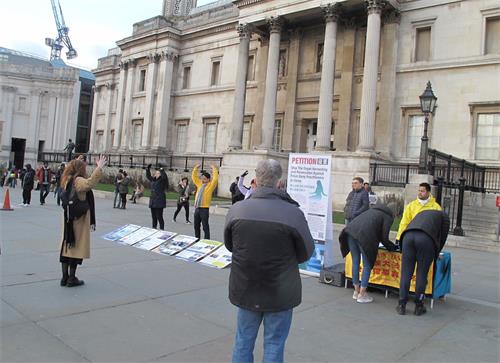 '图4:二零一九年二月二日,法轮功学员在伦敦特拉法加广场(Trafalgar Square)北平台的功法展示吸引过路民众现场学炼,图中一位西人女士正在模仿法轮功第二套功法“法轮桩法”中的“头顶抱轮”动作。'
