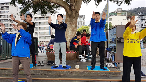 '图1:学员炼功,游客坐在树下观看真相展板。'