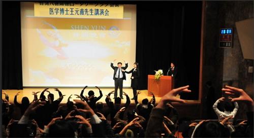 图4:二零一二年一月十四日,王元甫在东京新宿的会堂演讲“不花钱、不吃药的气功健康法”。图为,王元甫在讲台上演示法轮功第二套功法——法轮桩法,台下与会者跟着学炼动作。